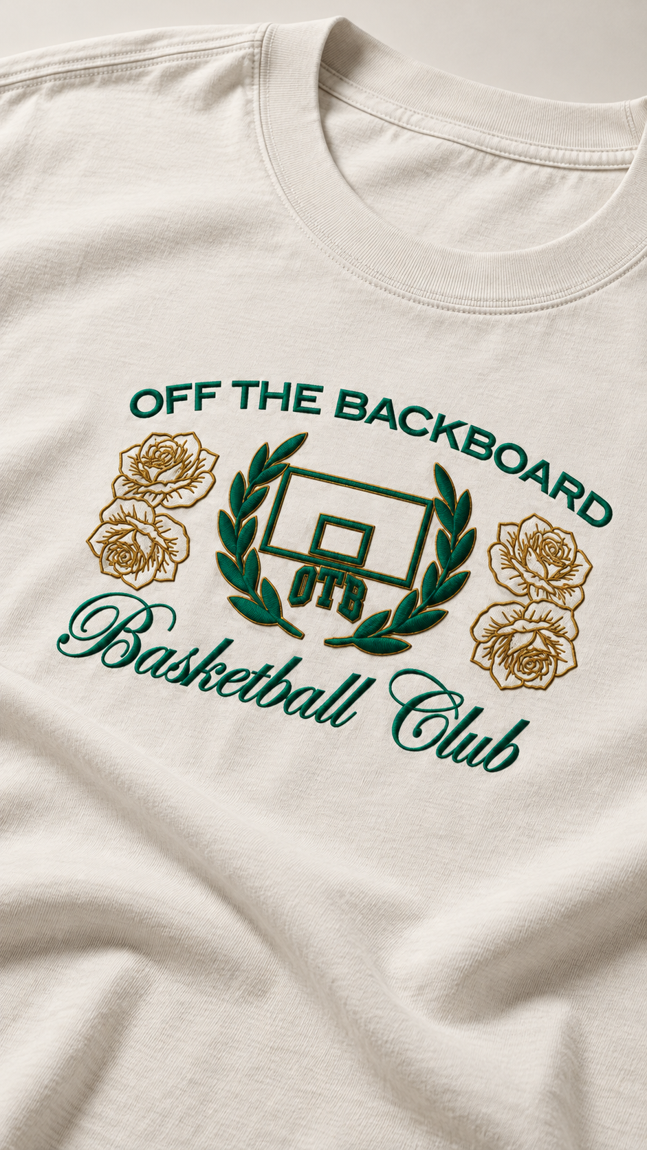 Close-up of a cream-colored t-shirt featuring a green and gold graphic that reads 'OFF THE BACKBOARD Basketball Club' with a basketball hoop, laurel wreath, and roses.