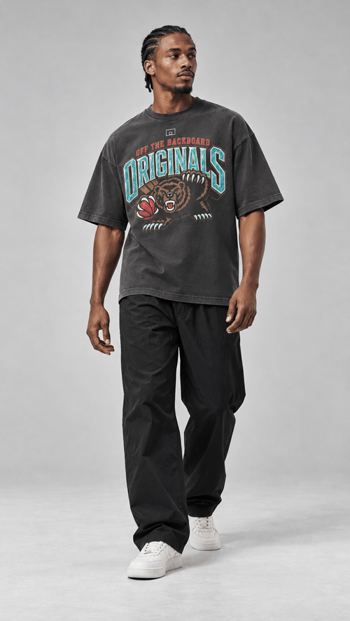 A man modeling a dark gray graphic t-shirt with 'ORIGINALS' and a bear logo, paired with black pants and white sneakers, against a light gray studio background.