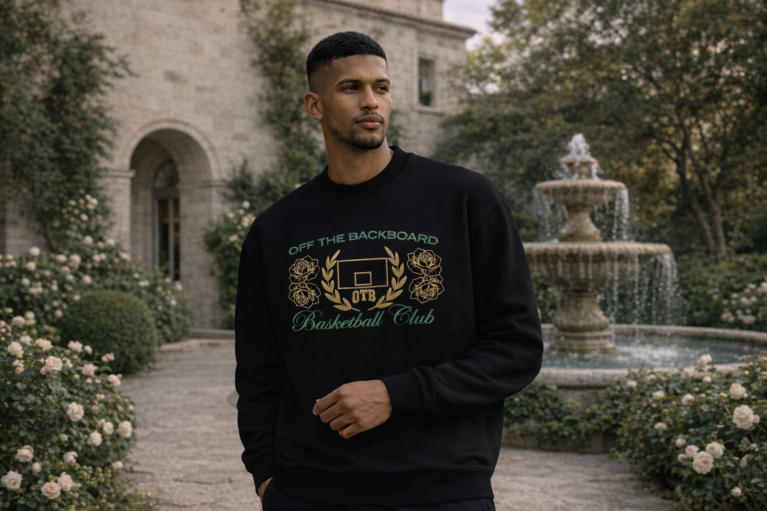 Man wearing a black sweatshirt with 'Off The Backboard Basketball Club' text and design, standing in a garden with a fountain.