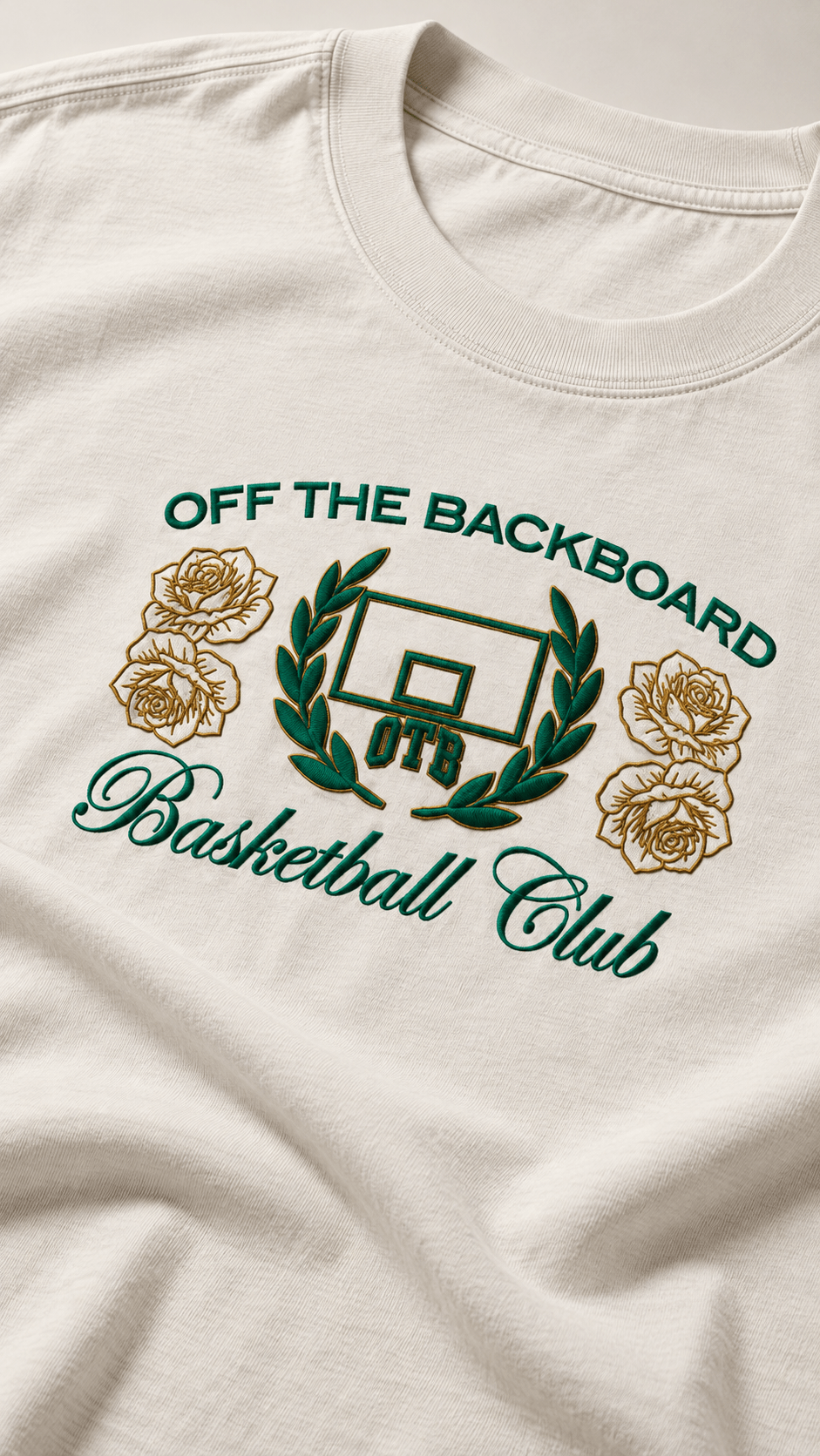 Close-up of a cream-colored t-shirt featuring a green and gold graphic that reads 'OFF THE BACKBOARD Basketball Club' with a basketball hoop, laurel wreath, and roses.