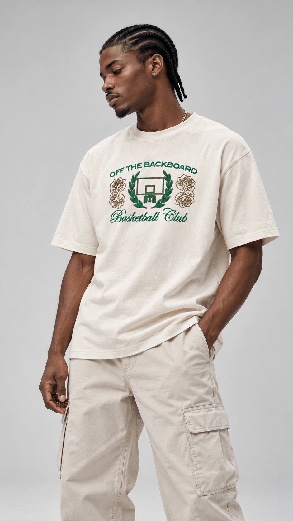 A man wearing a cream-colored graphic t-shirt with green and gold text reading 'OFF THE BACKBOARD Basketball Club' and matching cargo pants, photographed against a neutral gray studio background.